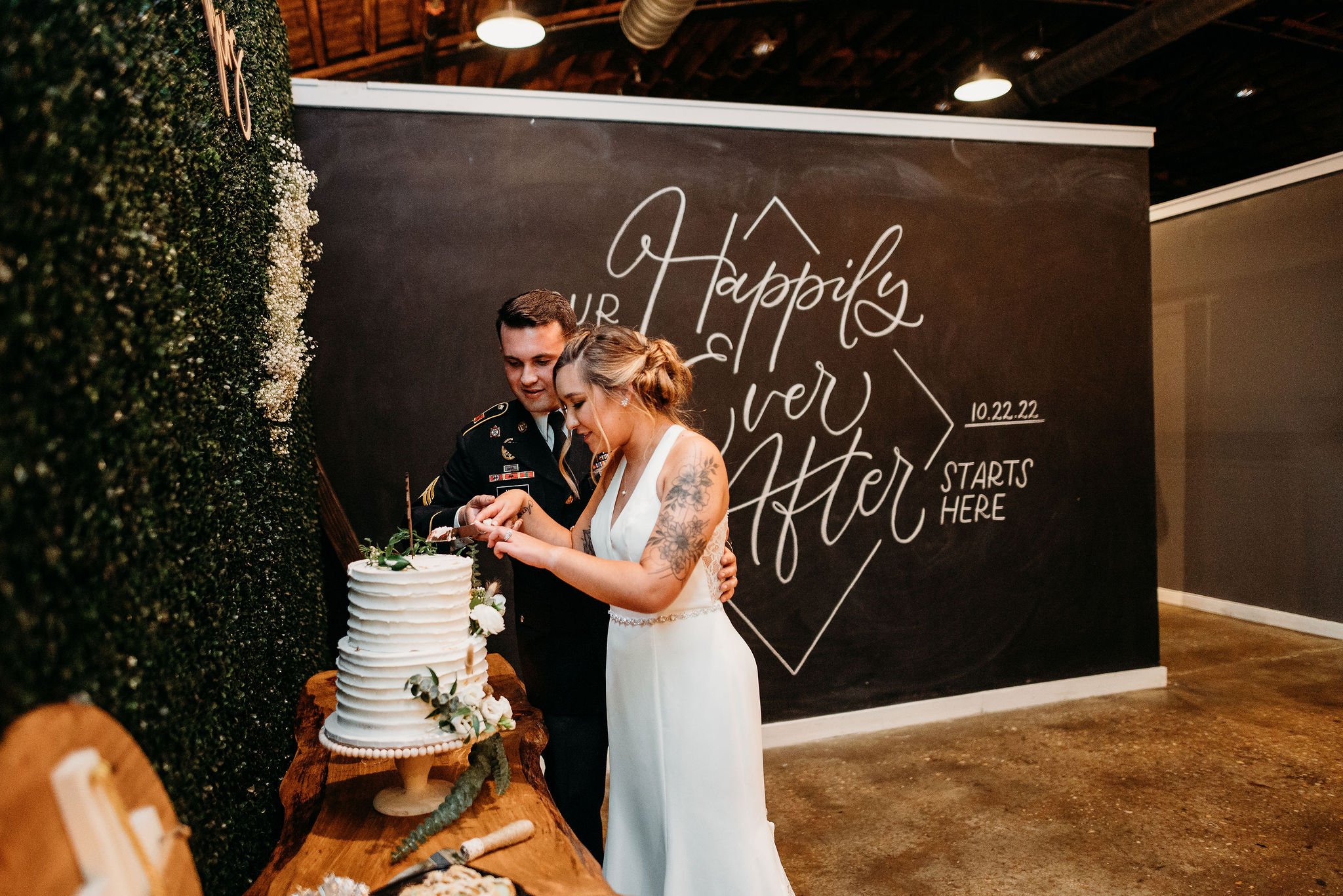A bride and groom cutting their wedding cake together in a rustic venue with a dark wall background that has a decorative sign reading 'Happily Ever After' and wedding date '10.22.22.'