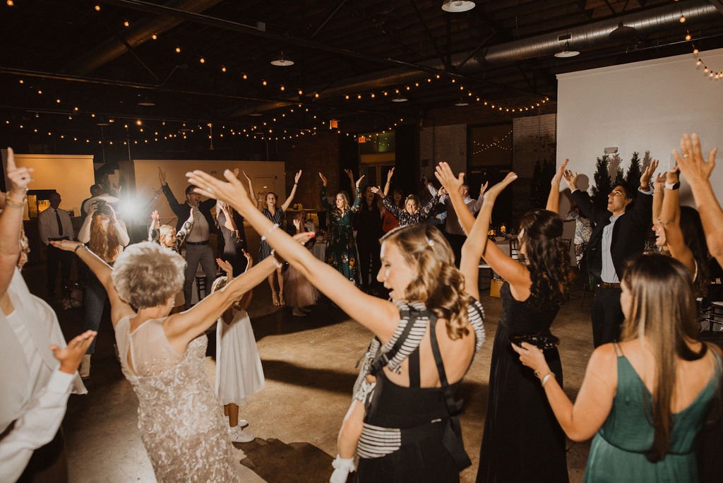 People dancing and celebrating at a wedding reception with string lights overhead.