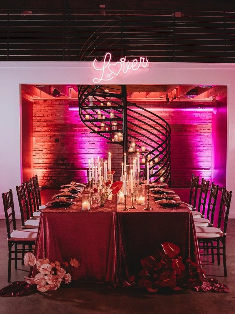 A romantic dining setup with a long table, candles, and floral decorations, illuminated with pink and purple lighting, under a neon sign that reads 'Lover', with a black spiral staircase and exposed brick wall in the background.