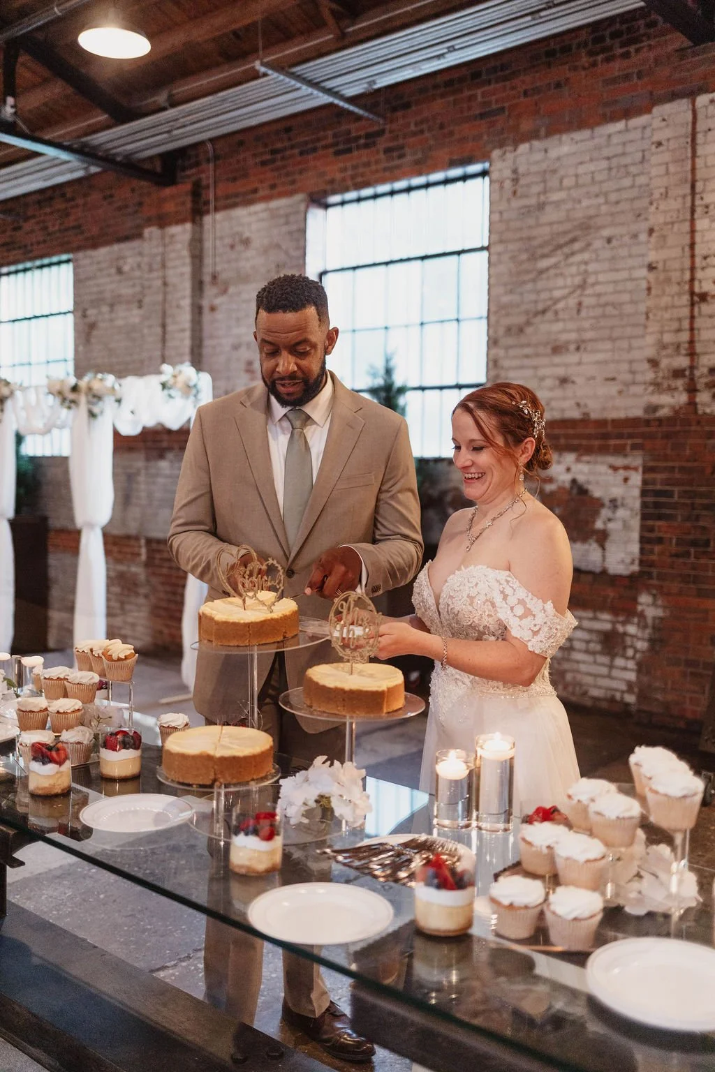 Bride and groom cutting their wedding cake at reception with cupcakes and desserts on table.
