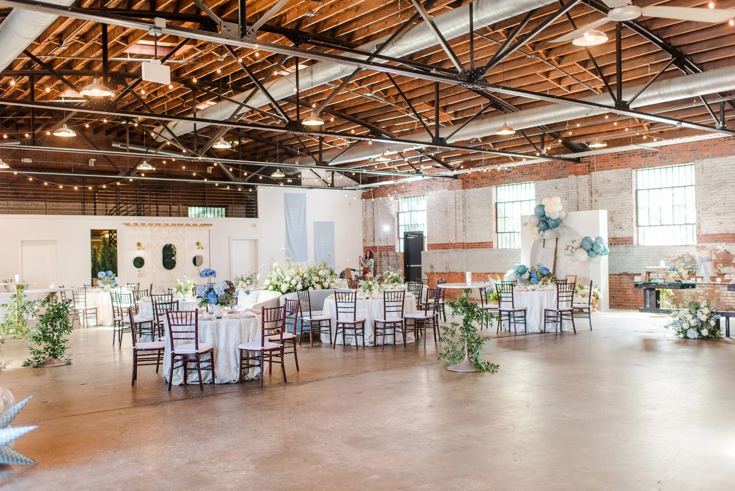 Large industrial-style event space decorated for a celebration with round tables covered in white tablecloths, floral centerpieces featuring white and blue flowers, and balloons arranged in clusters. The room has exposed brick and wooden beams on the ceiling, large windows allowing natural light, and some server stations or dessert tables along the walls.