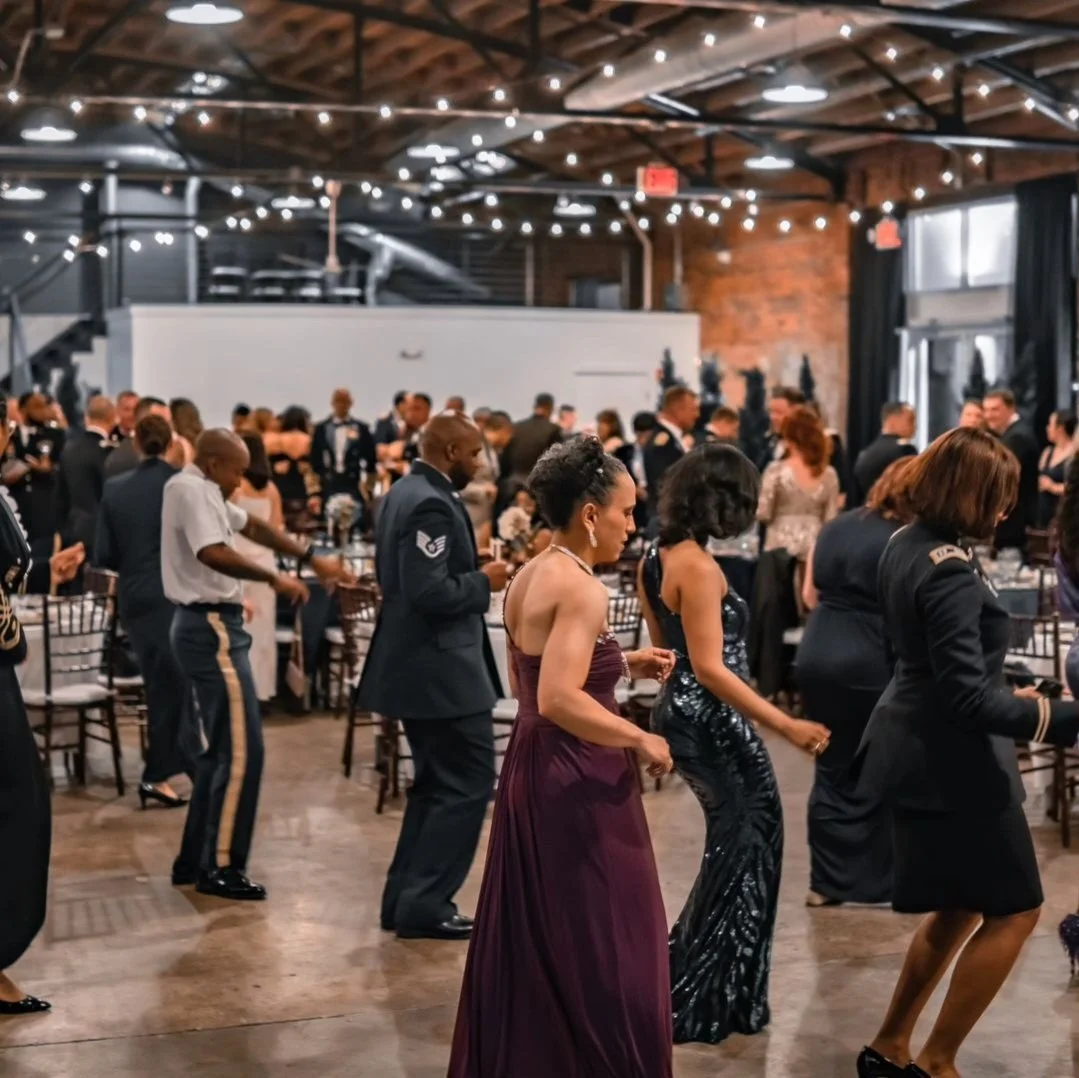 People dressed in formal attire are dancing at a wedding reception inside a decorated venue with string lights and wooden beams.