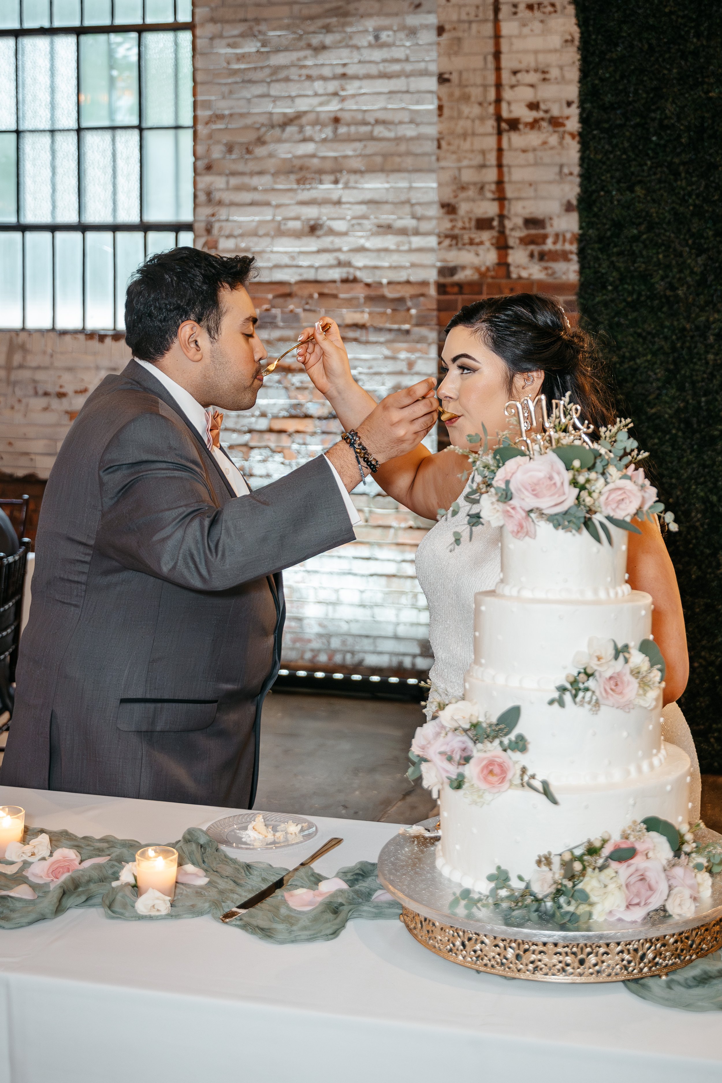 A bride and groom feed each other cake during their wedding reception. The wedding cake is decorated with pink and white flowers.