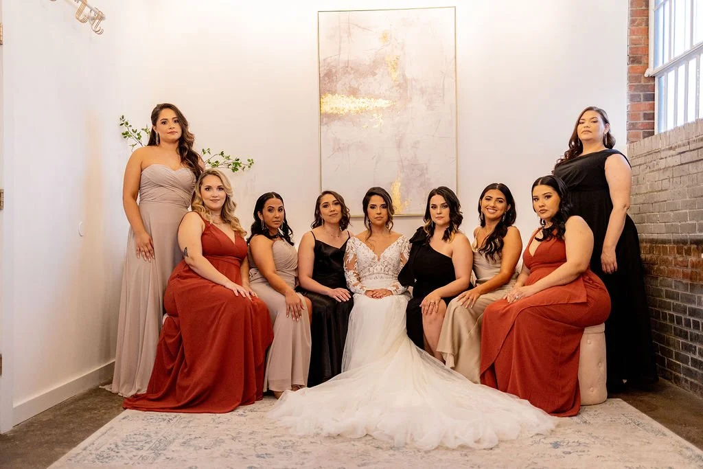 A group of ten women, including a bride in a white wedding gown, seated and standing in a modern indoor venue with white walls, brick accents, and large windows, posing for a wedding photo.