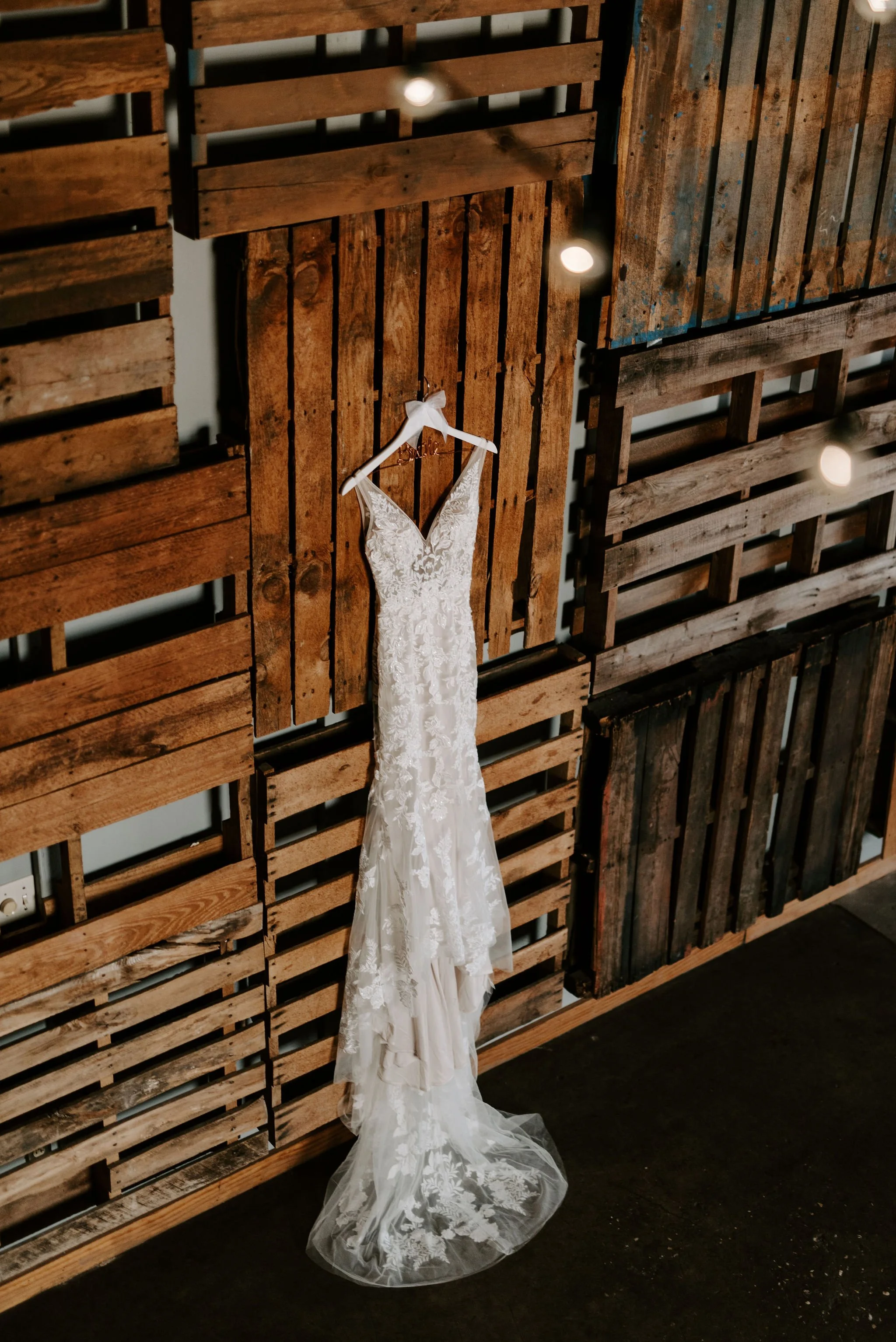 A white lace wedding dress hanging on a hanger against a rustic wooden wall.