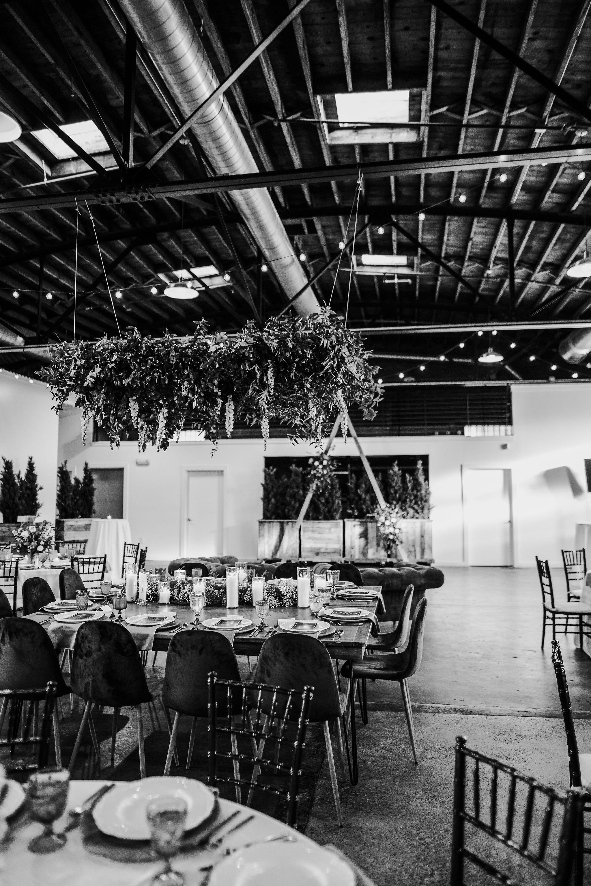 Black and white photo of an event space with decorated tables, hanging greenery centerpiece, and string lights on the ceiling.