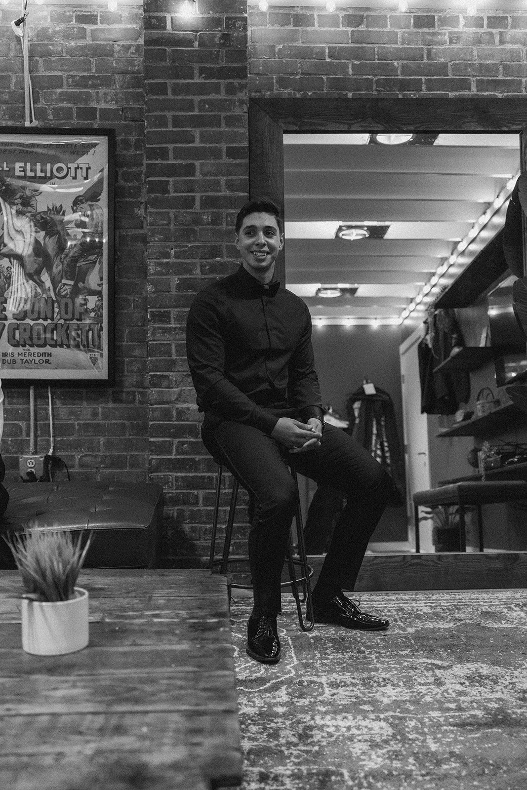 A man dressed in a black shirt and pants, wearing shiny black shoes, sitting on a high stool in a cozy, industrial-styled room with exposed brick walls, a framed poster to the left, and a doorway behind him leading to a room with shelves and equipment, in black and white.