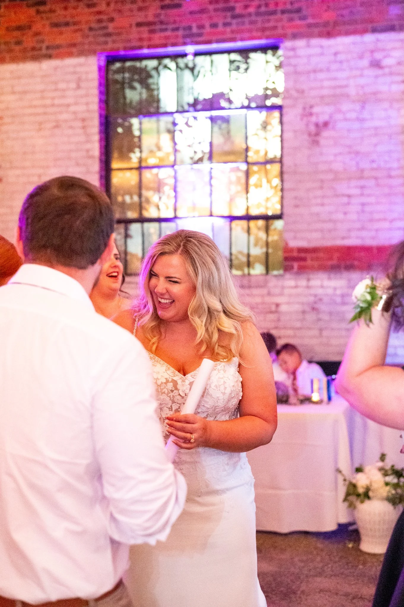 A woman in a white lace wedding dress holding a rolled document, smiling and laughing while interacting with a man in a white shirt at a wedding reception. The background features a brick wall, a multicolored window, and decorated tables.