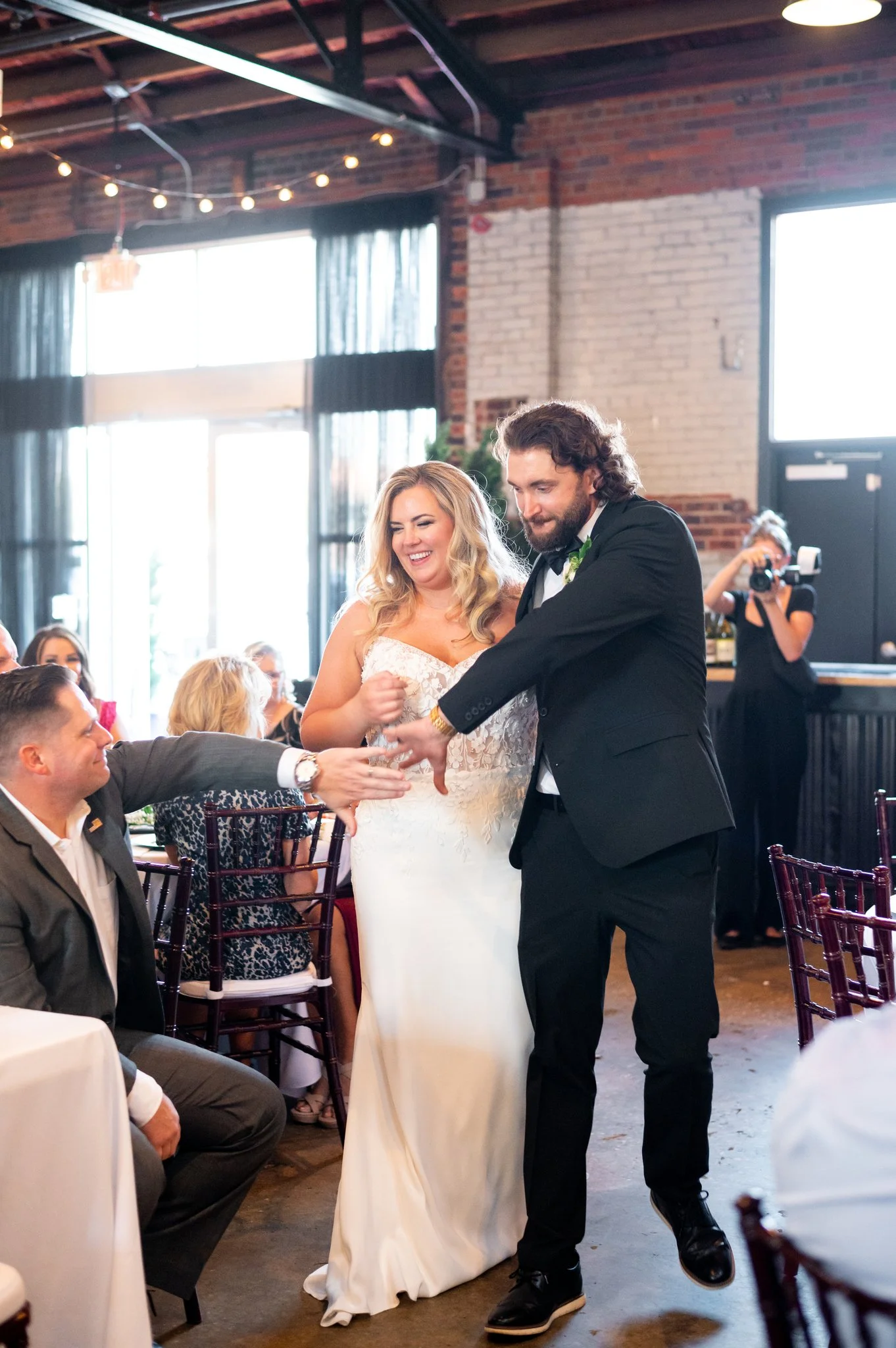 A bride and groom share a dance at their wedding reception, with guests watching and a photographer capturing the moment in a rustic indoor venue.