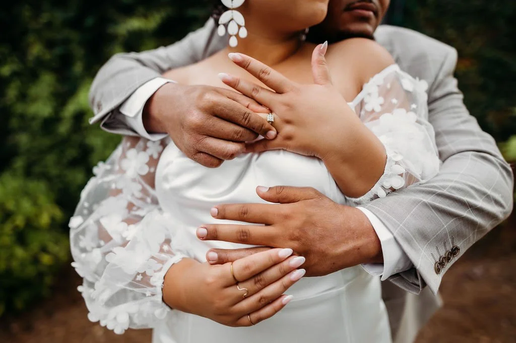 A couple embracing outdoors, with the woman in a wedding dress and the man in a light gray suit, showing close-up of their hands and arms.