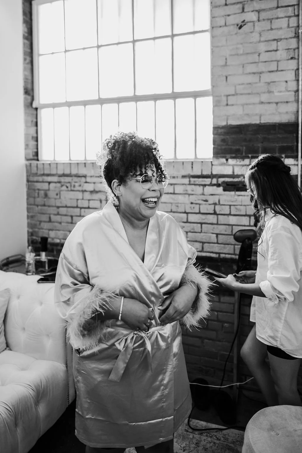 A woman in a silky robe is laughing, standing in a room with exposed brick walls and large windows, while another woman with hair covering her face holds a device nearby.