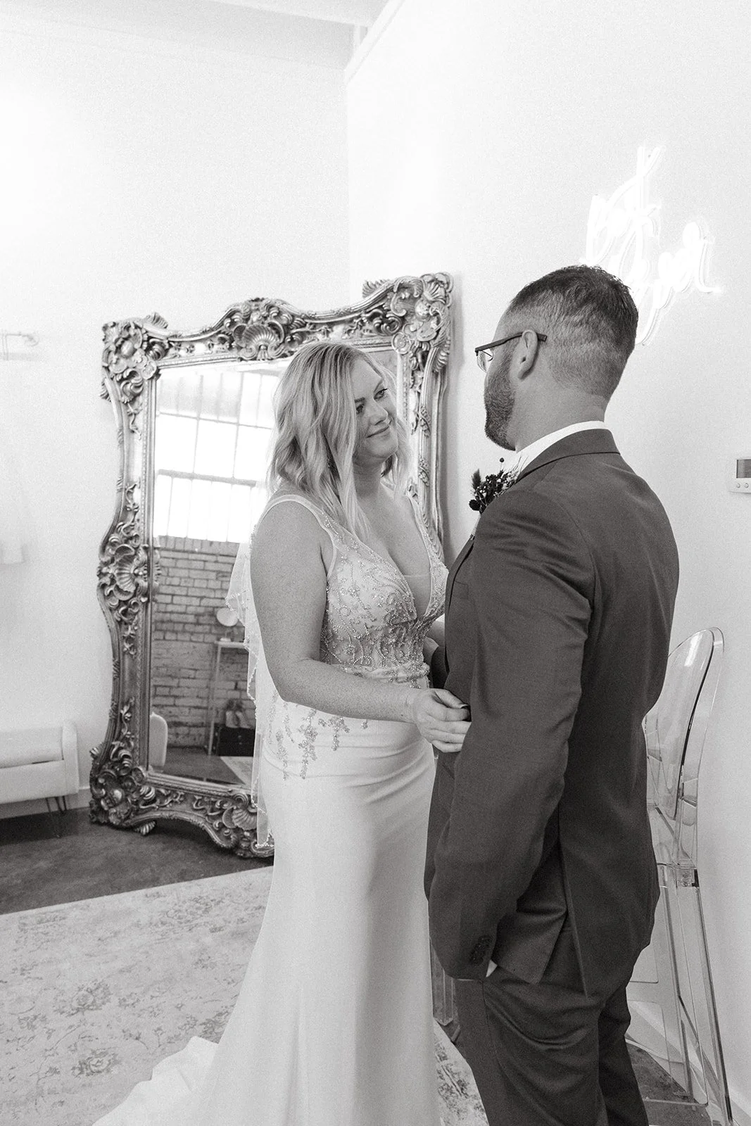 A bride and groom holding hands during their wedding ceremony indoors, with a large ornate mirror and a neon sign in the background.