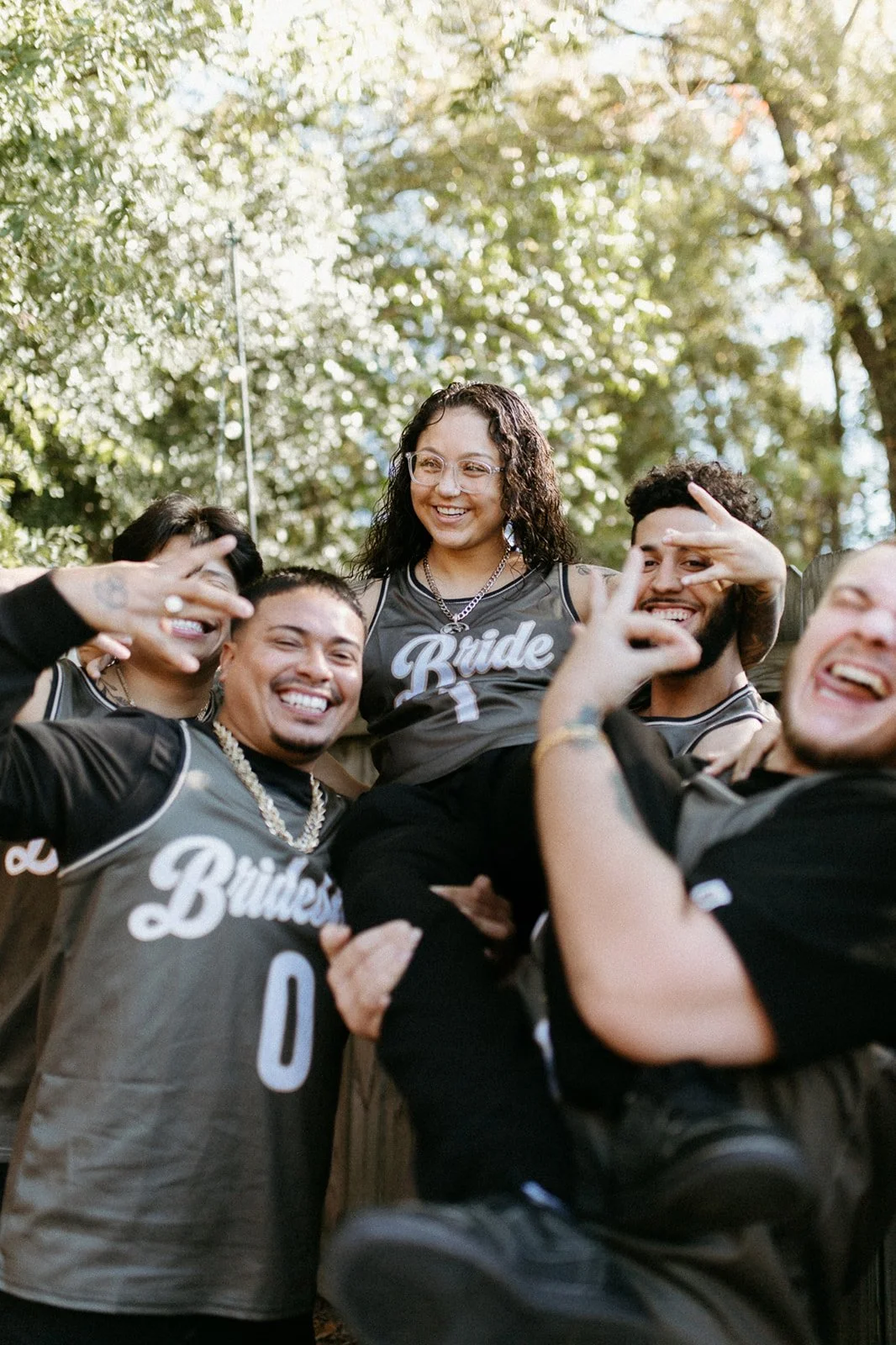 Group of five friends, some wearing black T-shirts with 'Brides' printed on them, smiling and holding each other outdoors with trees in the background, celebrating a wedding.