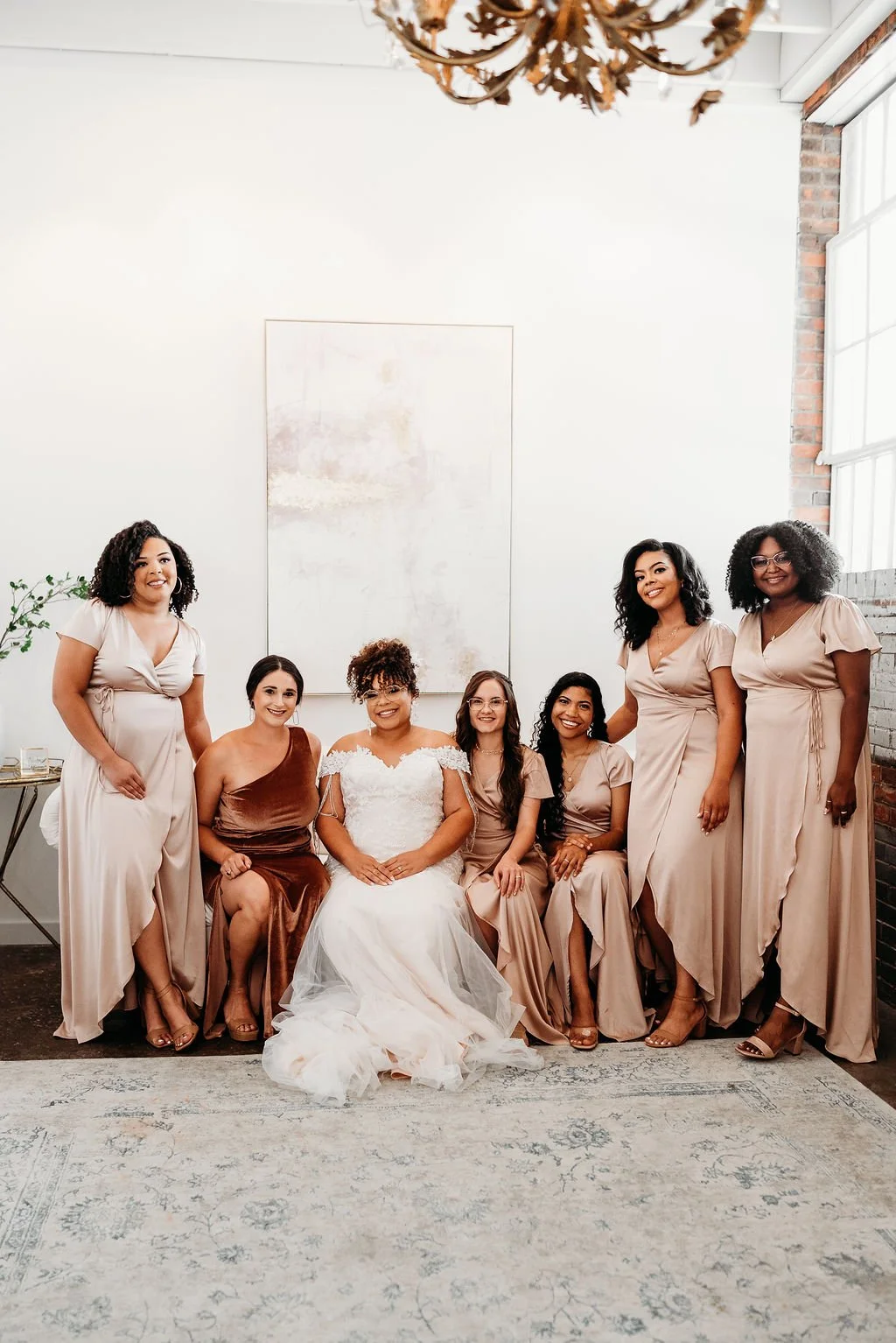 A bride with six women, all dressed in coordinated beige and brown gowns, standing and sitting in a bright, modern room with brick and white walls, a large abstract painting, and a chandelier.