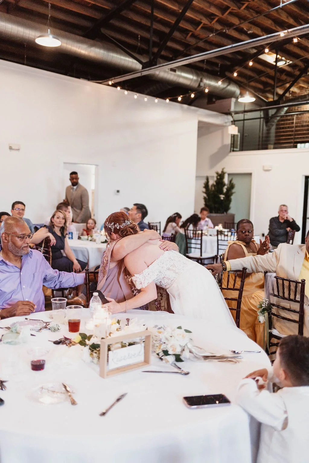 A wedding reception scene where a bride is hugging an older woman at a round table decorated with flowers and candles, with other guests seated around the table in a spacious venue with industrial ceiling and string lights.