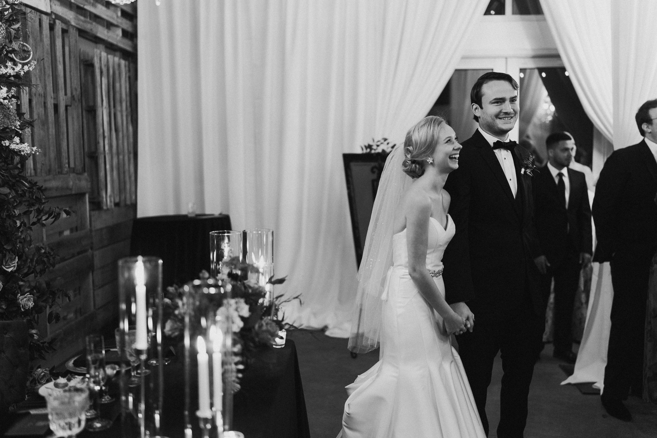 A bride and groom holding hands and smiling during their wedding reception, with other guests in the background, decorated with candles and flowers.