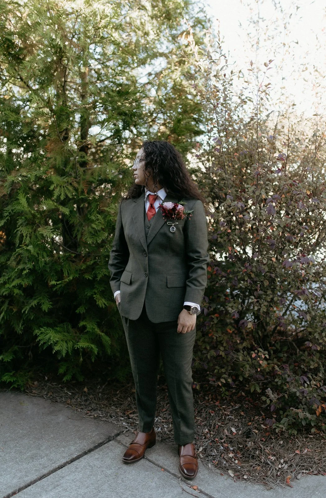 A woman in a dark gray business suit with a red tie and white shirt, standing outdoors on a sidewalk near green and purple bushes, with sunlight filtering through the trees, wearing glasses and a wristwatch, with a boutonnière pinned to her lapel.