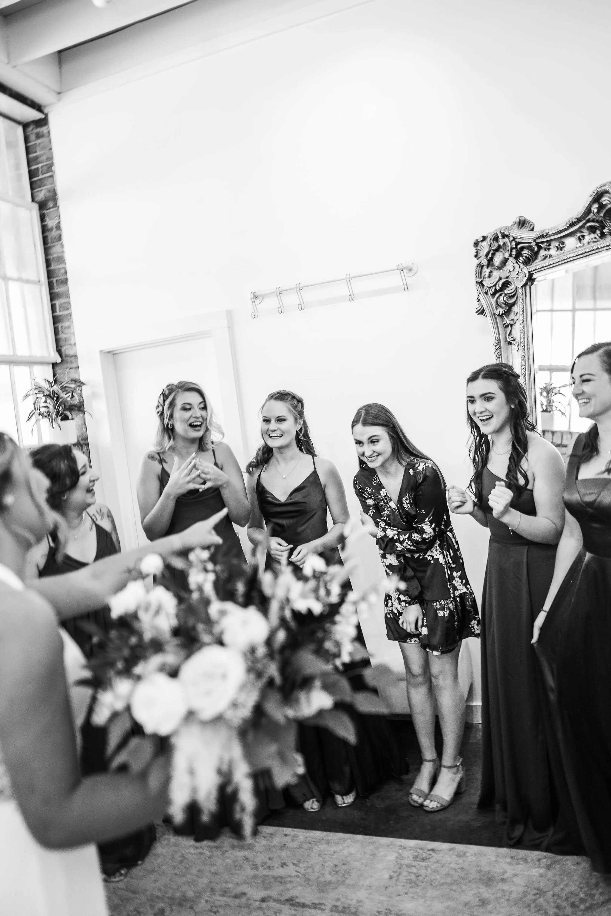 Group of women in formal dresses laughing and chatting in a bridal party room, with a woman holding a bouquet in the foreground.