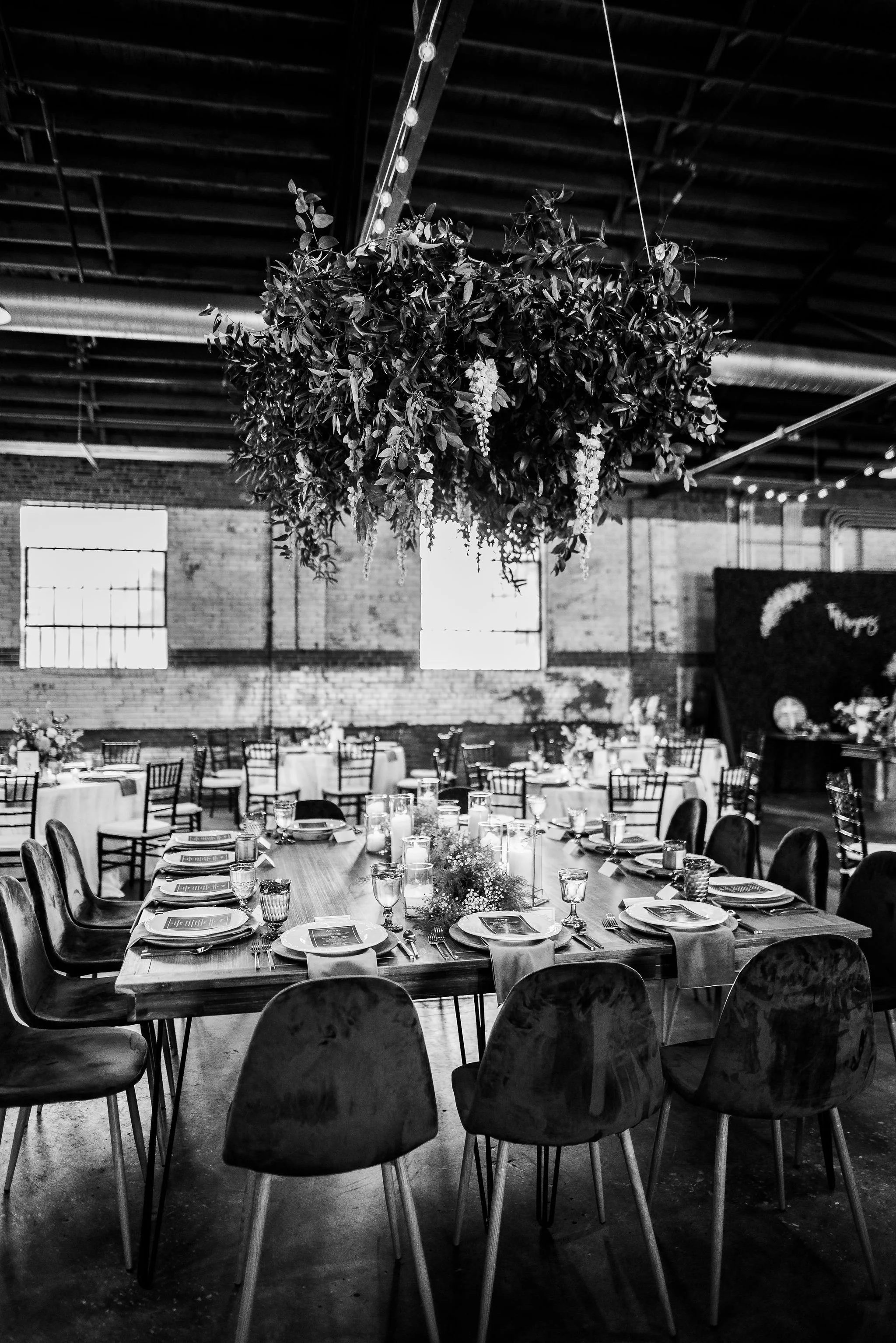 Black and white photo of a decorated event space with a large hanging floral arrangement, round tables with place settings, and a rustic industrial interior.
