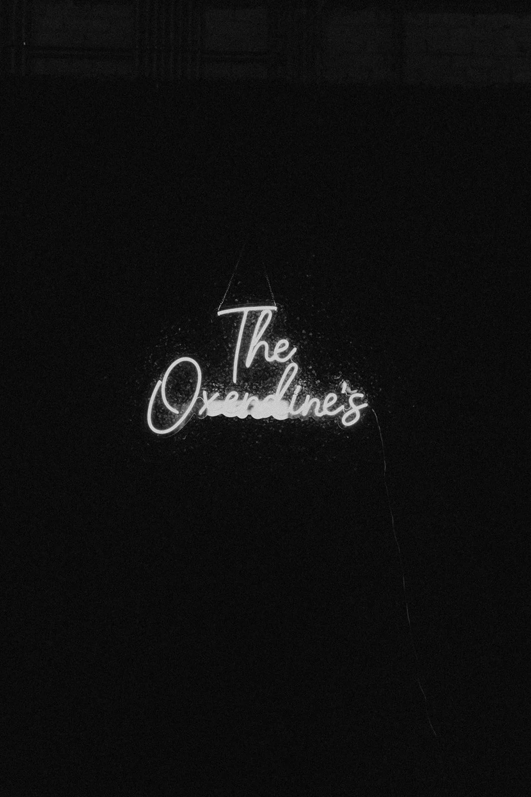 Neon sign that reads "The Overdue's" in cursive font, hanging on a dark wall.