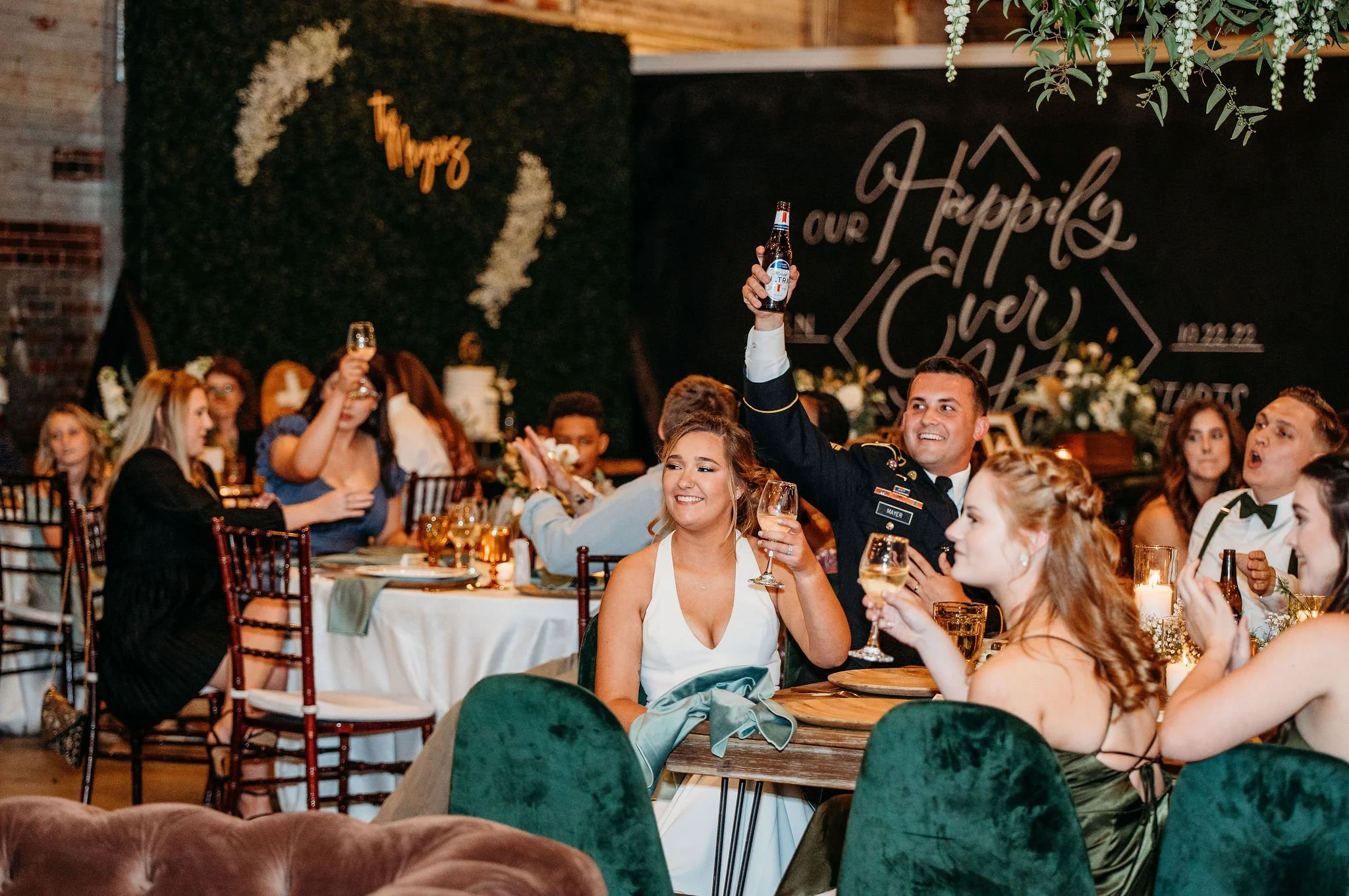 People celebrating at a wedding reception, raising glasses of drinks and smiling, with a decorated backdrop and floral arrangements.