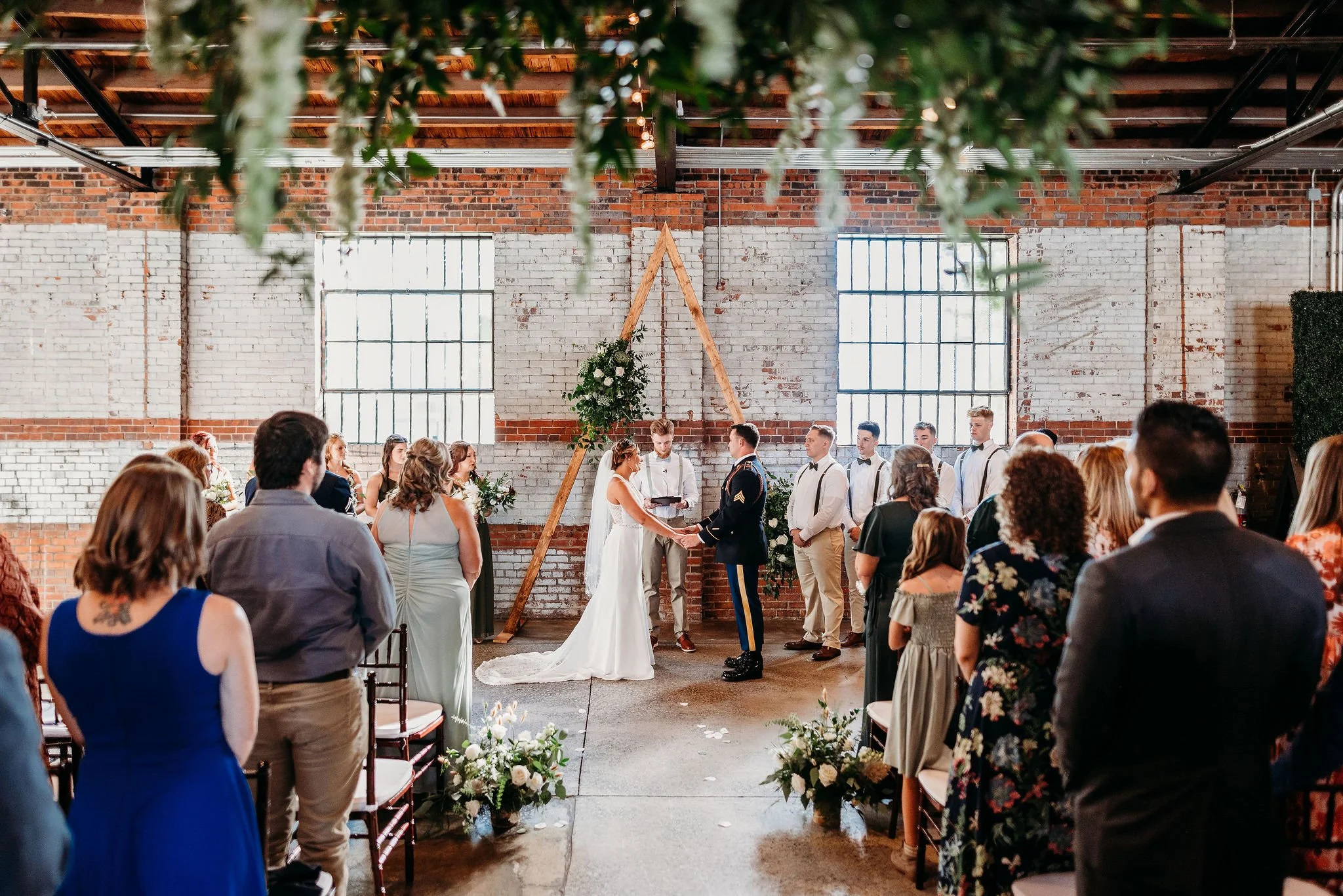 A wedding ceremony with a bride and groom holding hands in front of officiant, facing each other, inside an industrial-style venue with brick walls and large windows, decorated with greenery and floral arrangements, surrounded by seated guests.