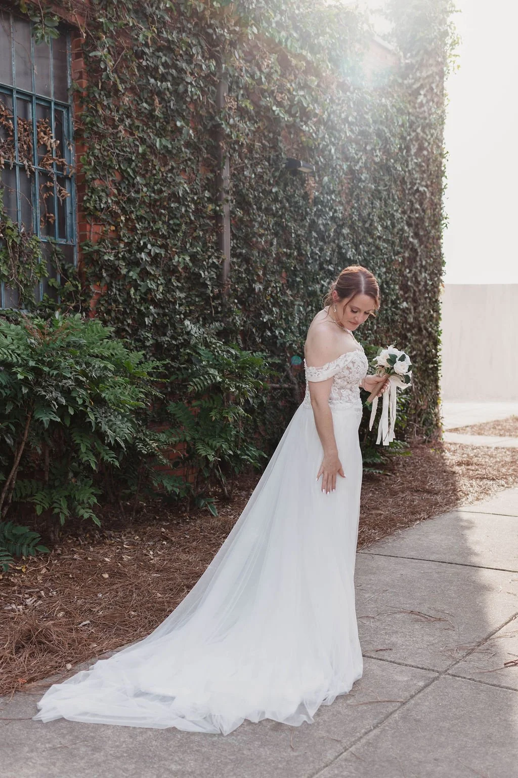 A bride in a white wedding dress holding a bouquet, standing outdoors near a brick wall covered in ivy on a sunny day.
