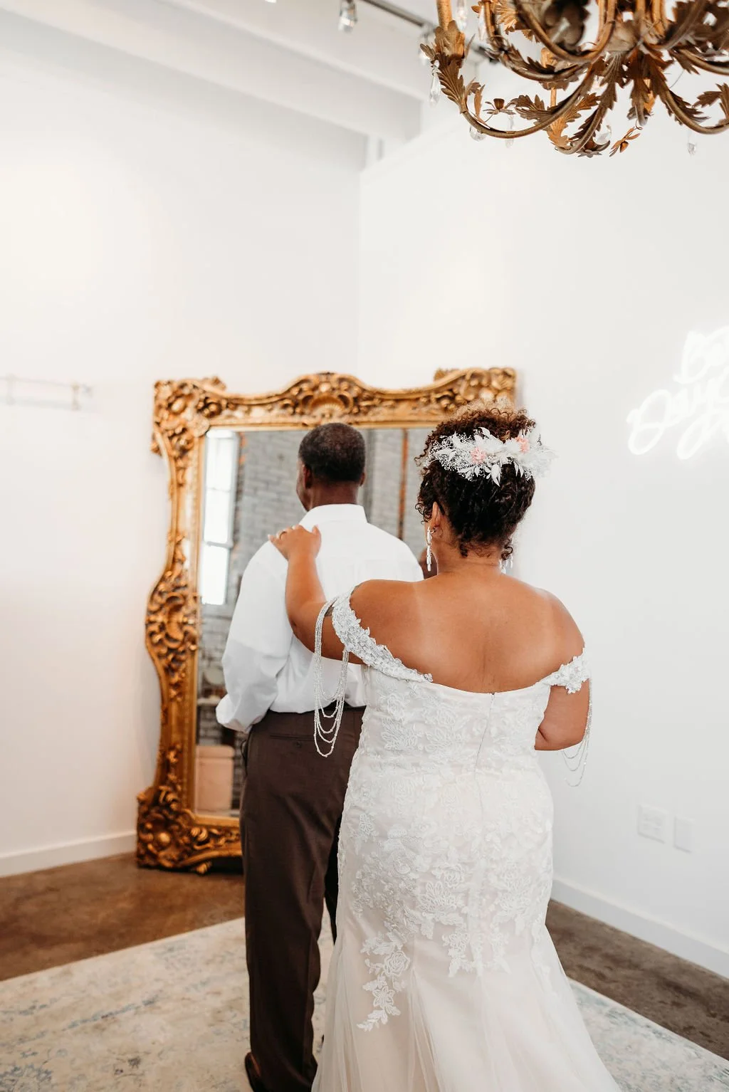 A bride with short curly hair and a floral headpiece, wearing an off-shoulder lace wedding gown, is standing behind a groom in a white dress shirt and dark trousers, touching his shoulder, as they look into a large ornate mirror in a decorated indoor