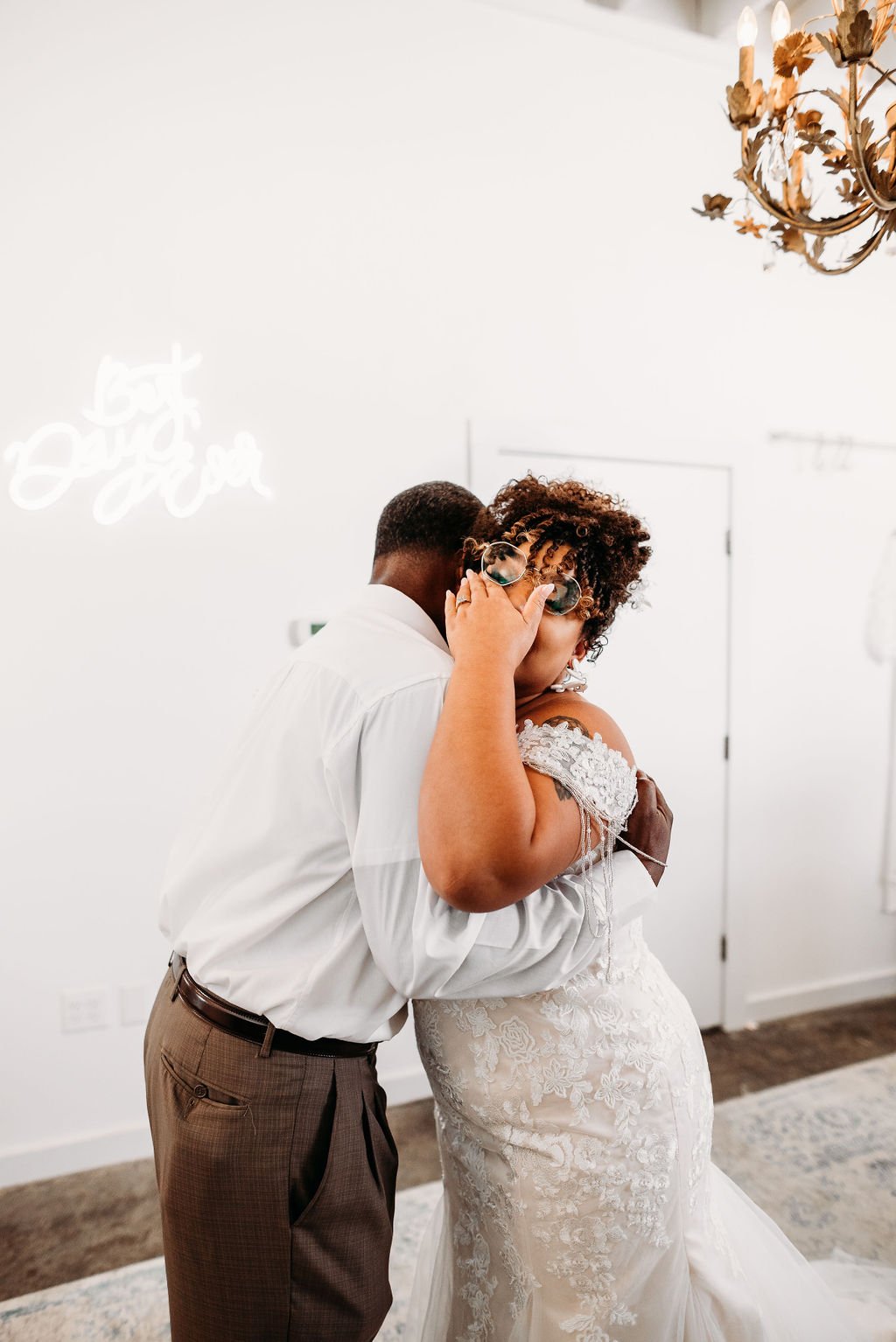 A couple sharing an intimate moment, with the person on the left wrapping their arms around the other. The person on the right is wearing a white lace wedding dress and glasses, holding the glasses with one hand while looking at the camera. The setti