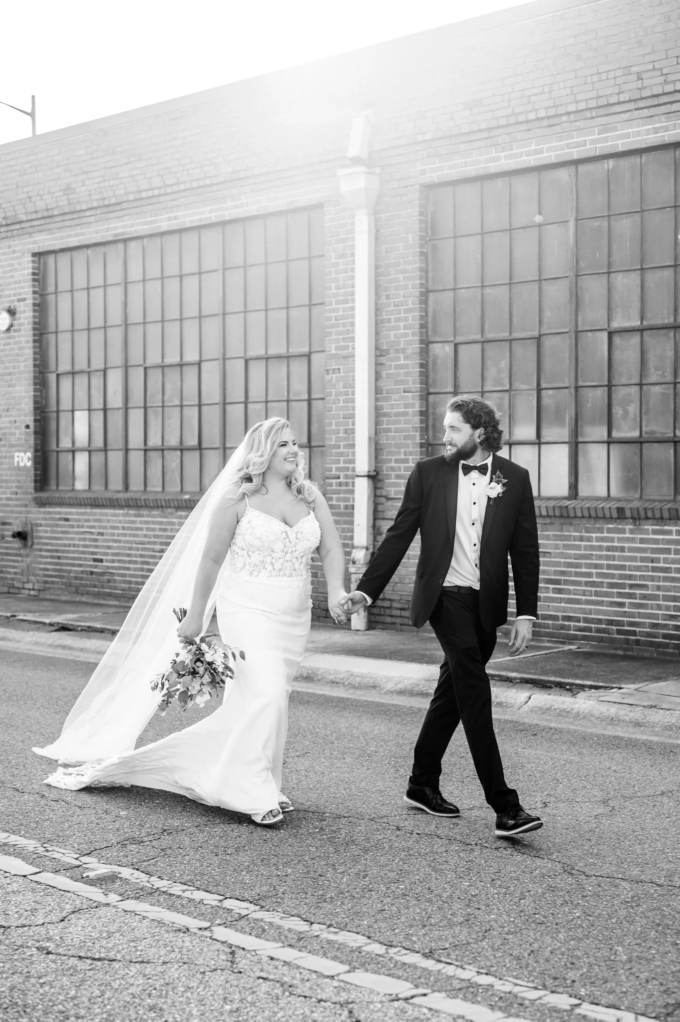 A bride and groom walking hand-in-hand on a city street, smiling at each other, with a brick building and large windows in the background.