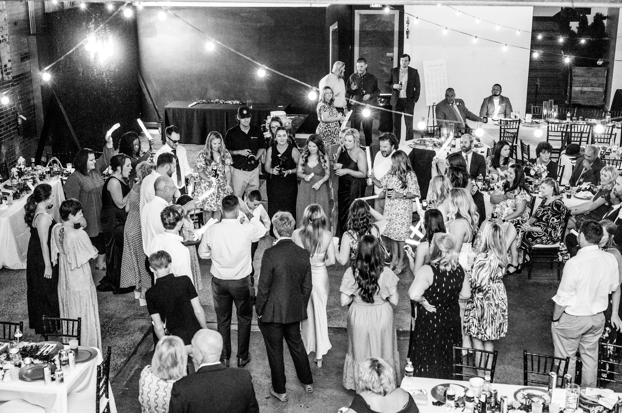 Black and white photo of a social gathering, with people dancing, chatting, and sitting at decorated tables with food and drinks in an indoor venue decorated with string lights.