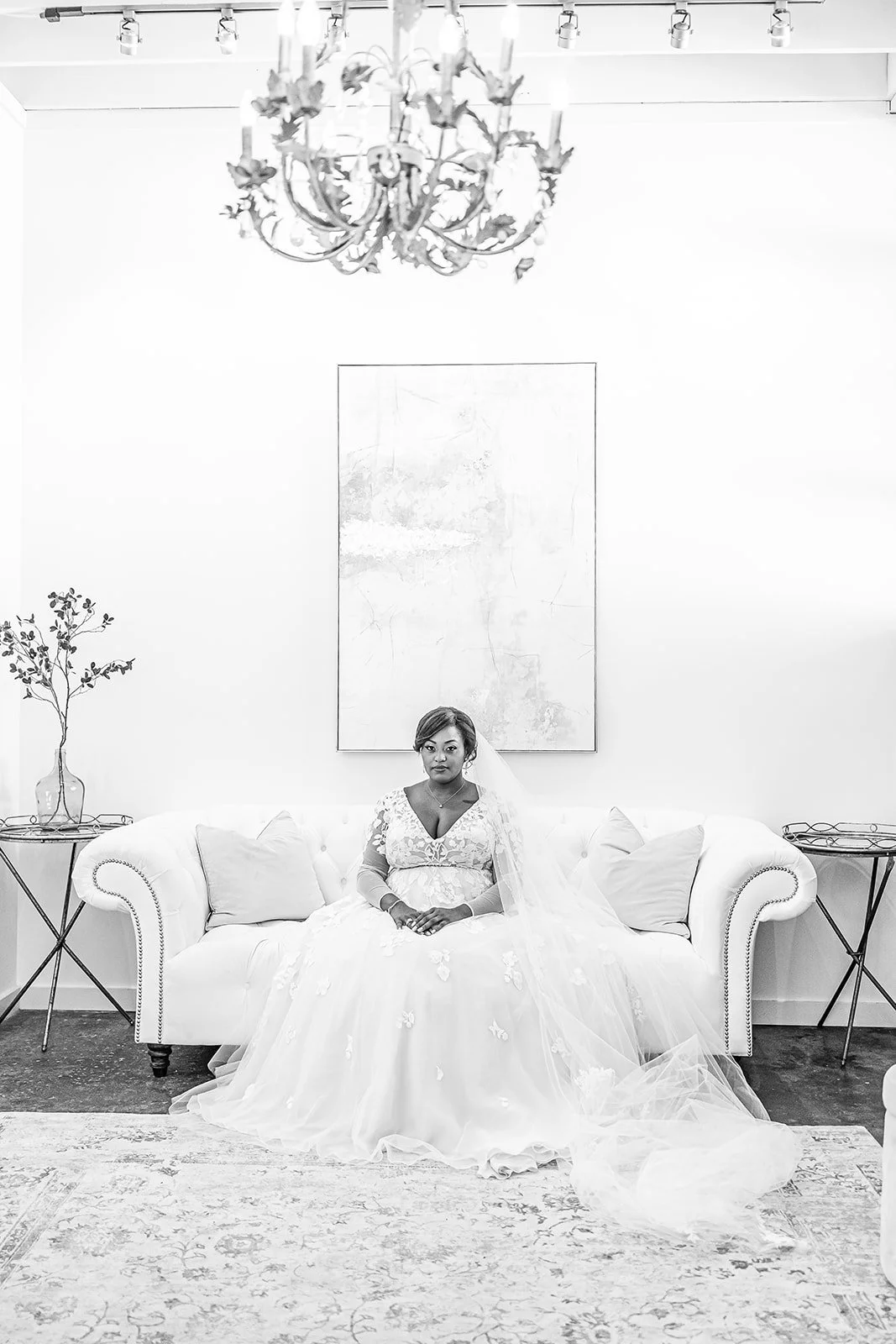 A woman in a wedding dress sitting on a white sofa in a room with a chandelier and decorative plants.