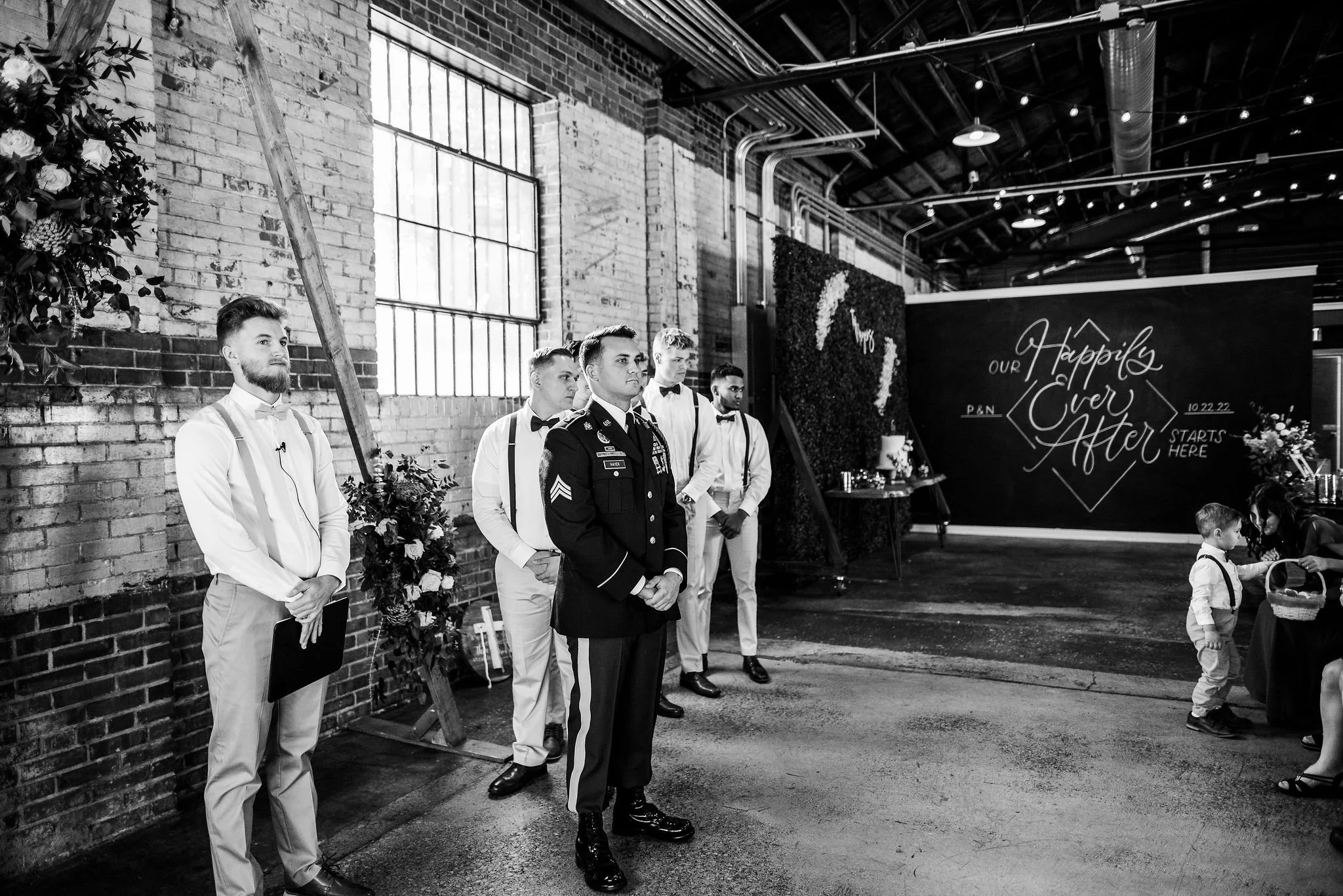 A black and white photo of a wedding reception with five men standing against a brick wall. One man in the center is dressed in a military uniform, and the others are in formal white shirts with suspenders or black bow ties. There is a large window o