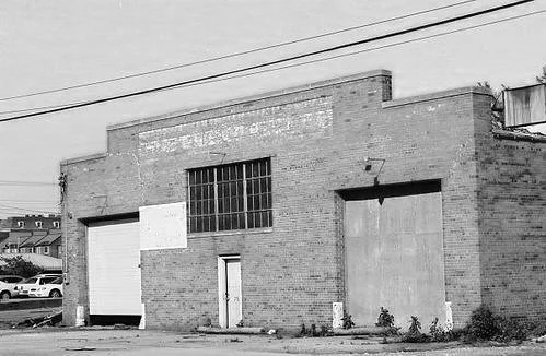 Abandoned brick building with large garage doors and a small door in the center, in an industrial area.