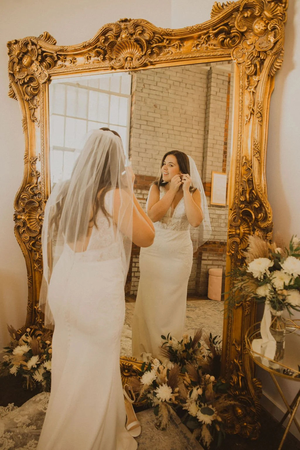 A woman in a wedding dress and veil is standing in front of a large ornate gold mirror, adjusting her earrings and smiling at her reflection.