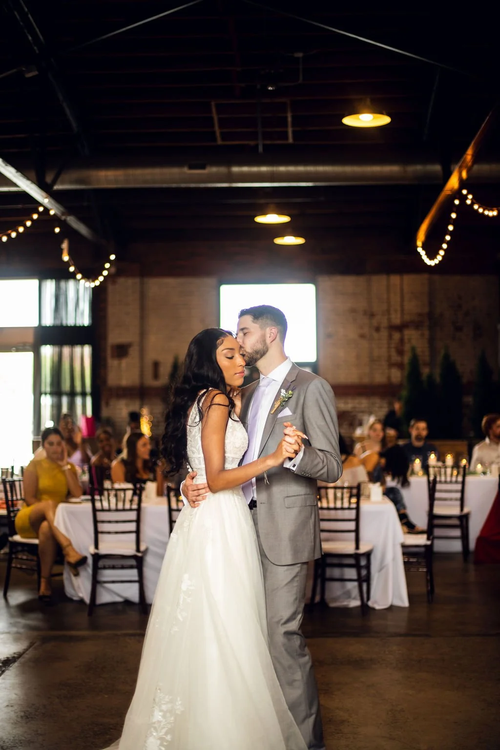 A bride and groom are dancing together at their wedding reception, with guests seated at decorated tables in the background.