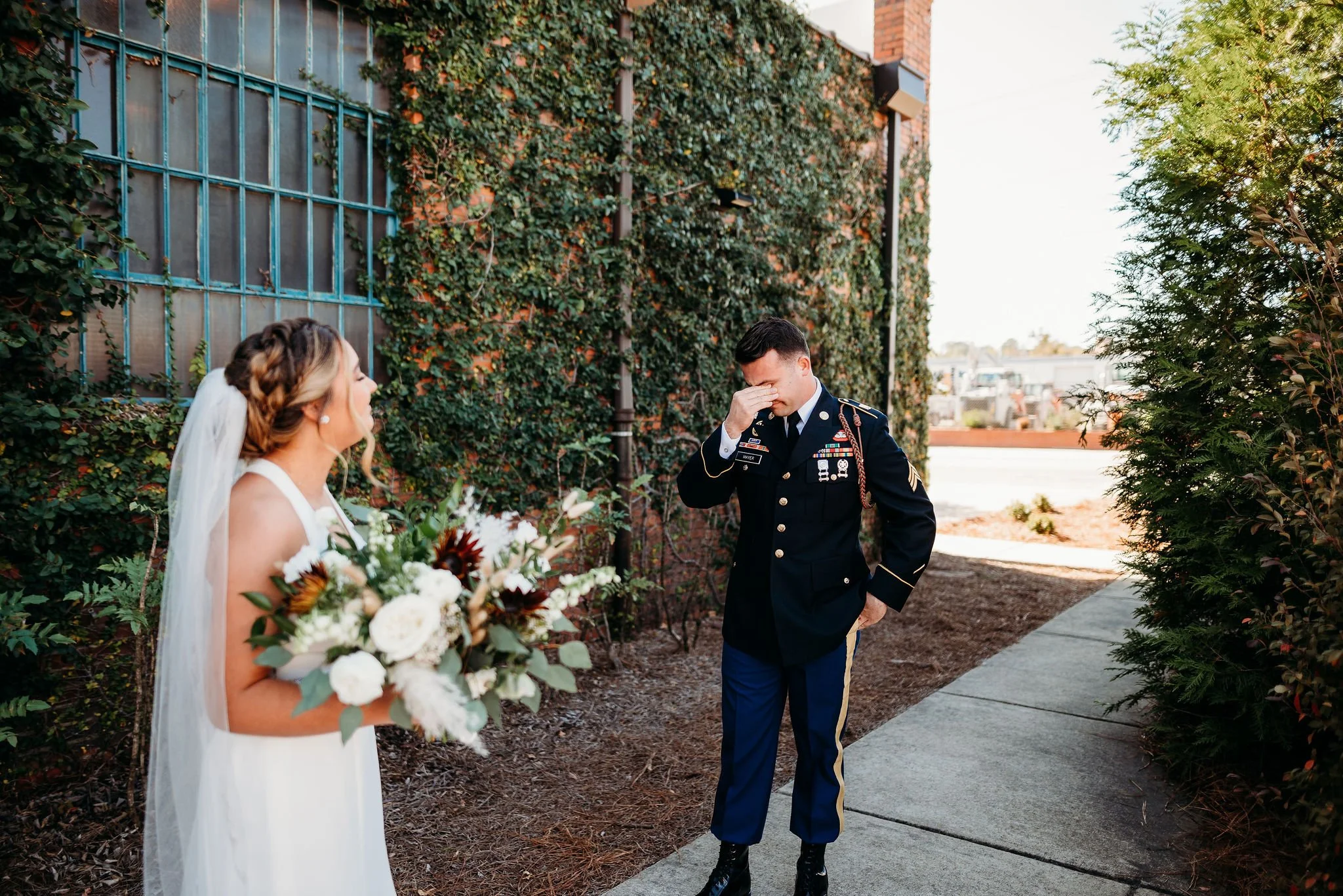 A bride holding a bouquet of flowers, standing outside and crying, while a man in a U.S. military uniform stands with his hand covering his face, appearing emotional.