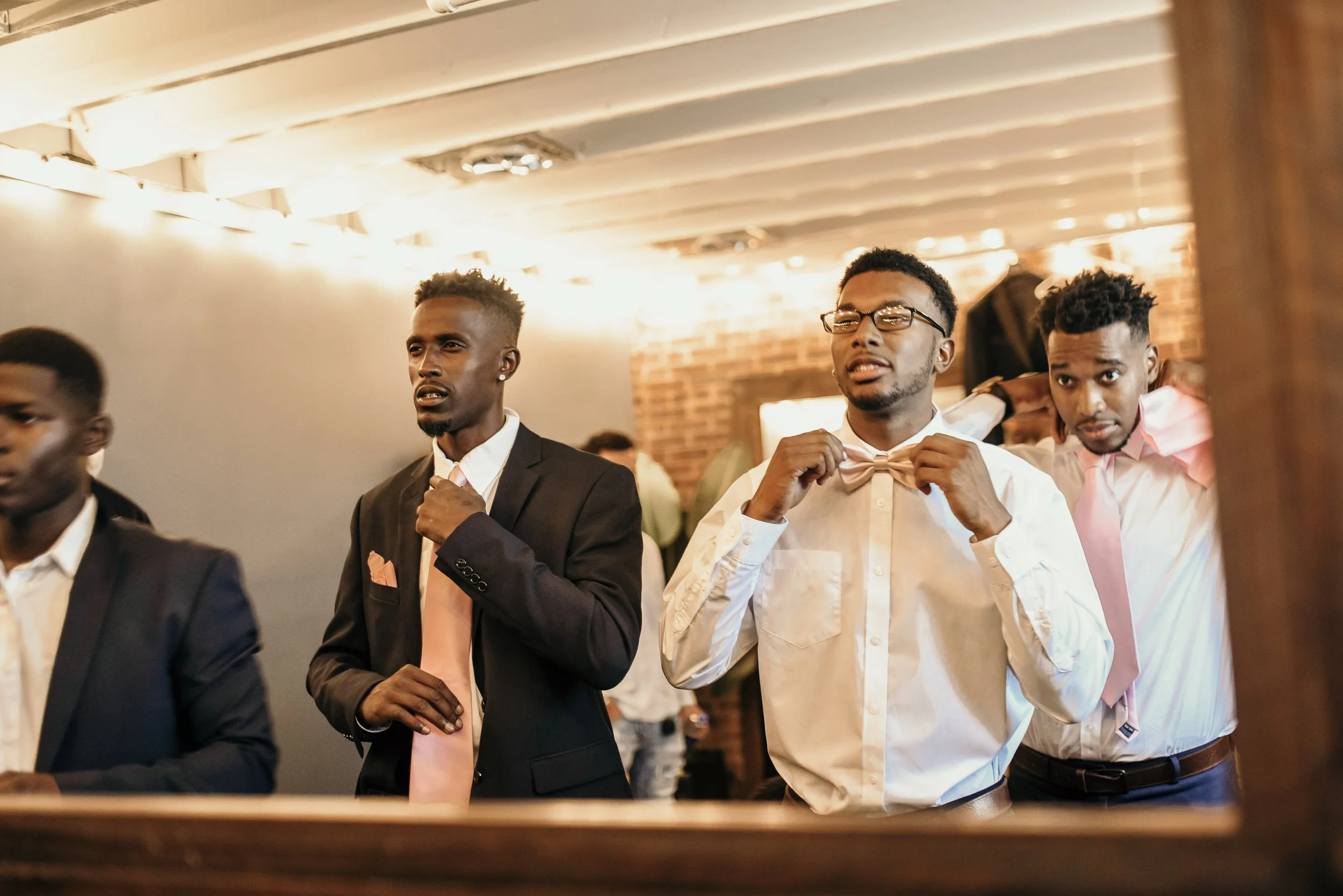 Three men dressed in formal attire, adjusting their neckties or bow ties, in an indoor setting with brick and white walls and warm lighting.