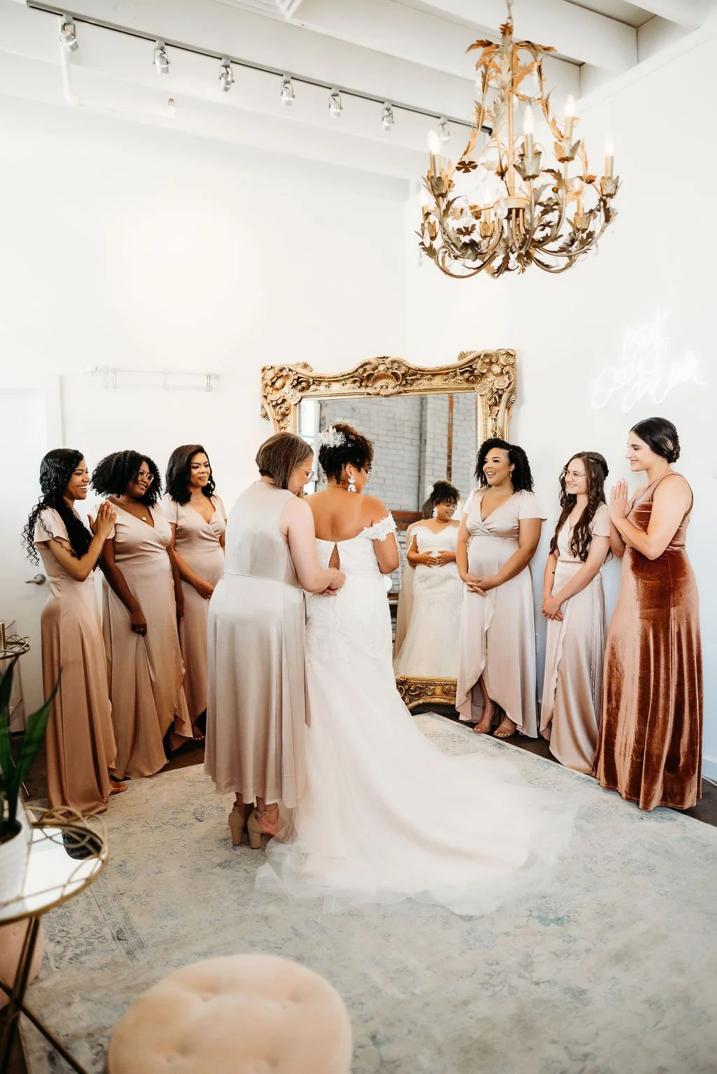 Bridal party with bride and bridesmaids in a room with a large ornate mirror and a chandelier, sharing a joyful moment before the wedding.