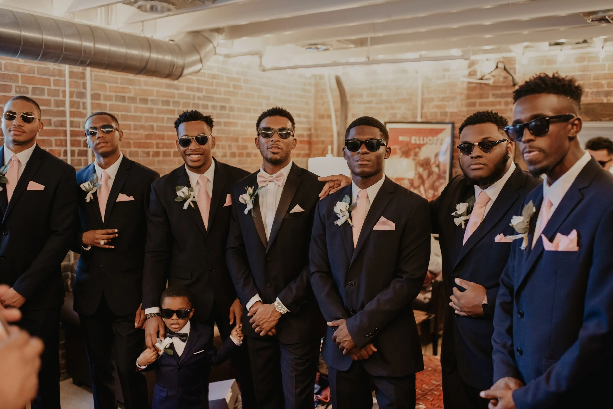 A group of eight men and a young boy dressed in formal suits with sunglasses, standing indoors with a brick wall background at a wedding or special event.