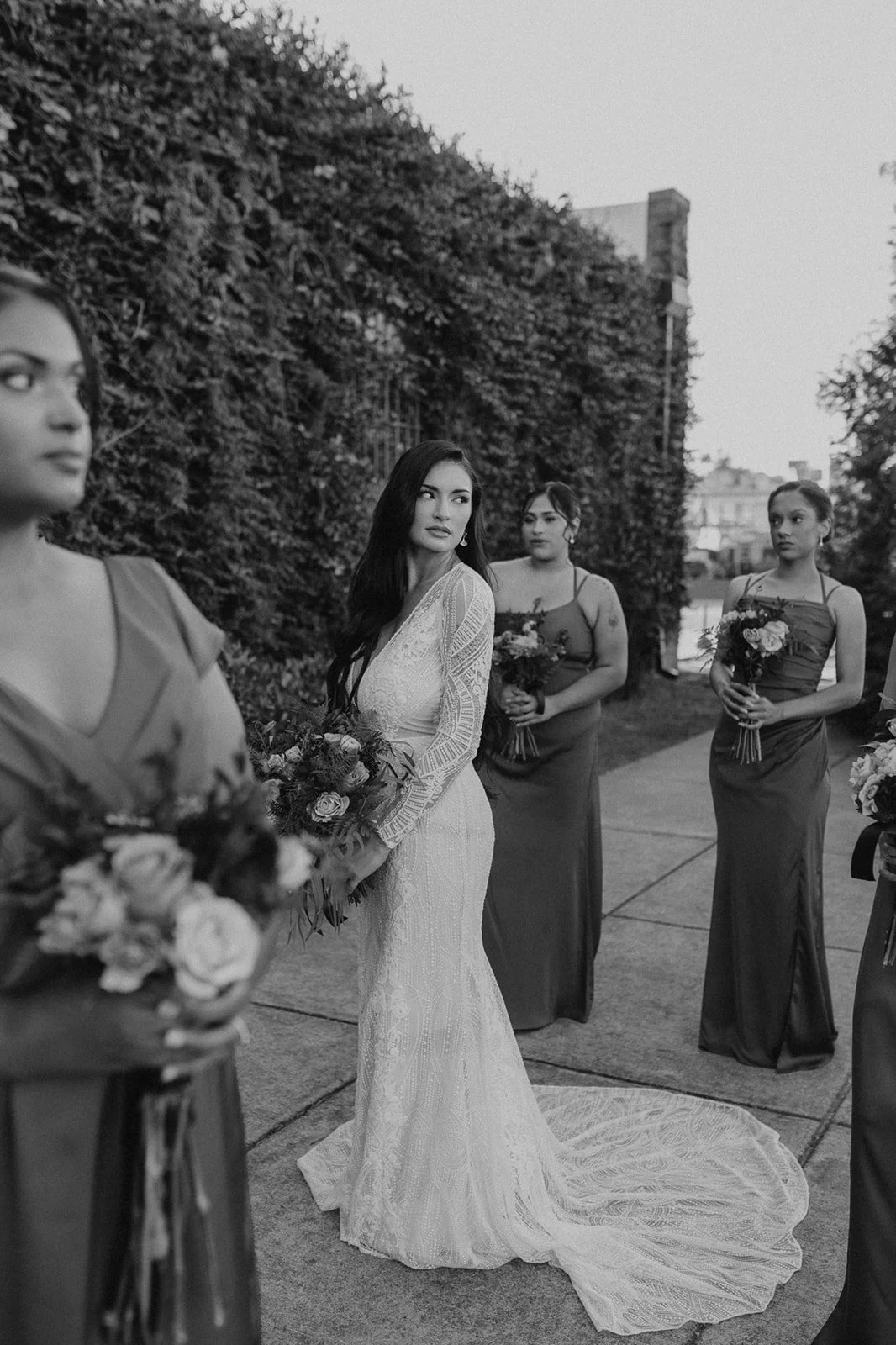 A black and white photo of a bride in a wedding dress with long dark hair holding a bouquet, standing outside with three bridesmaids in matching dark dresses also holding bouquets, against a background of trees and buildings.