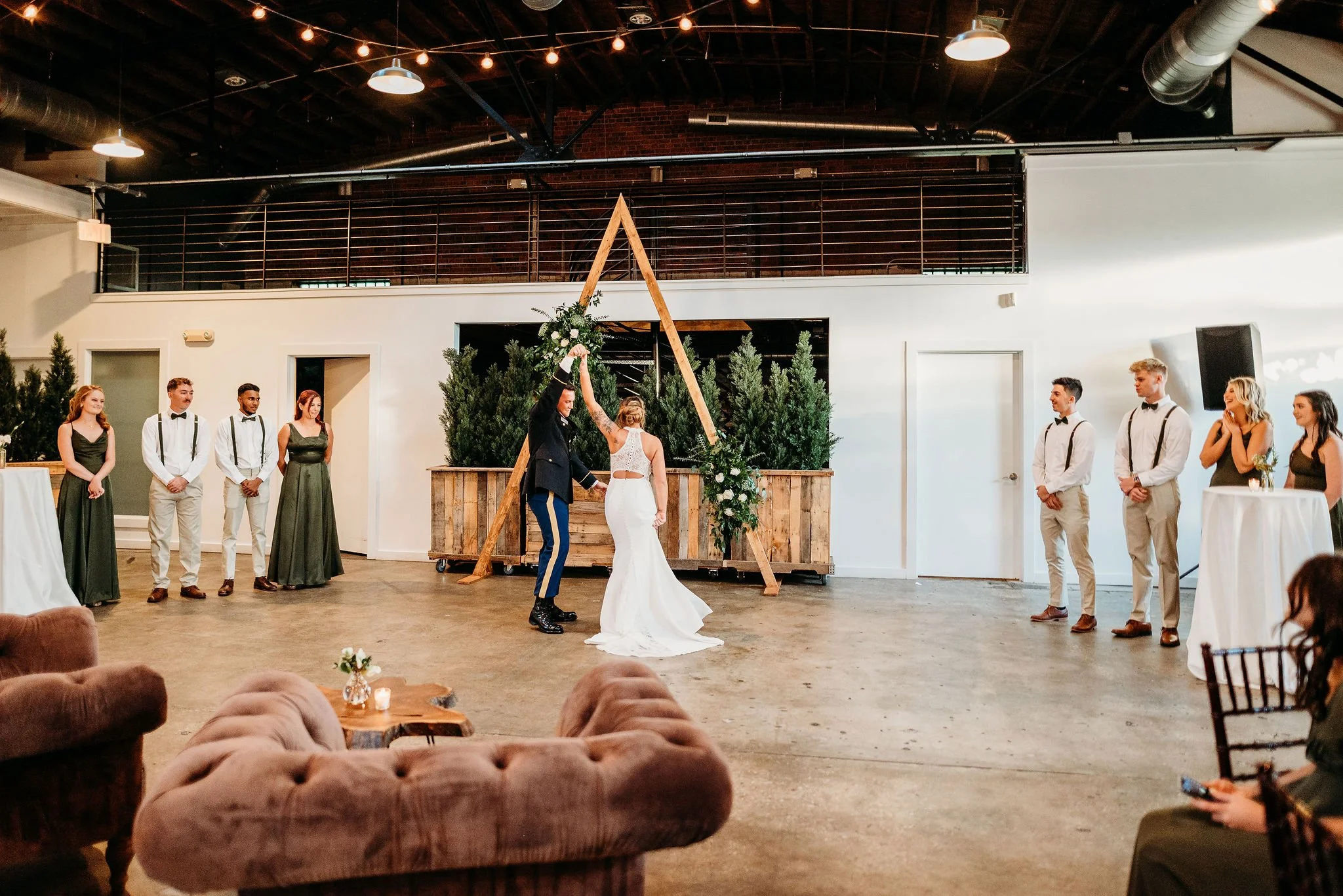 A wedding ceremony is taking place indoors with a bride and groom under a wooden arch decorated with flowers. The bride is wearing a white gown, and the groom is in a blue suit. Four women and four men are standing on either side, dressed in formal a
