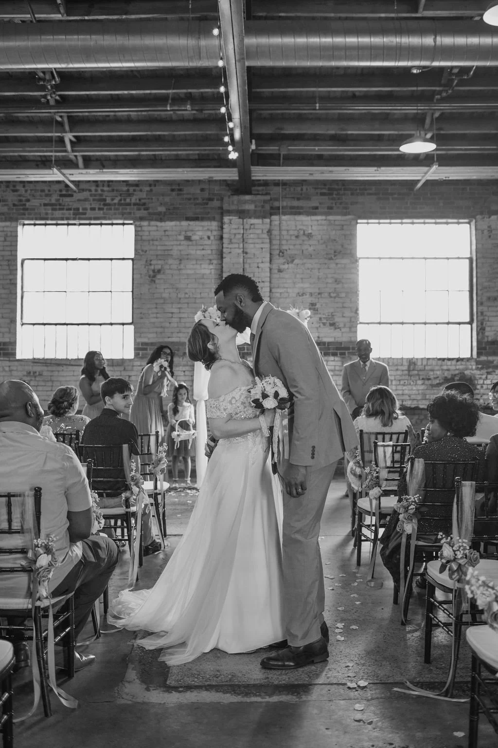 A black and white photo of a wedding ceremony in an industrial-style venue with brick walls and large windows. The bride and groom are sharing a kiss at the altar, surrounded by seated guests and flower decorations.