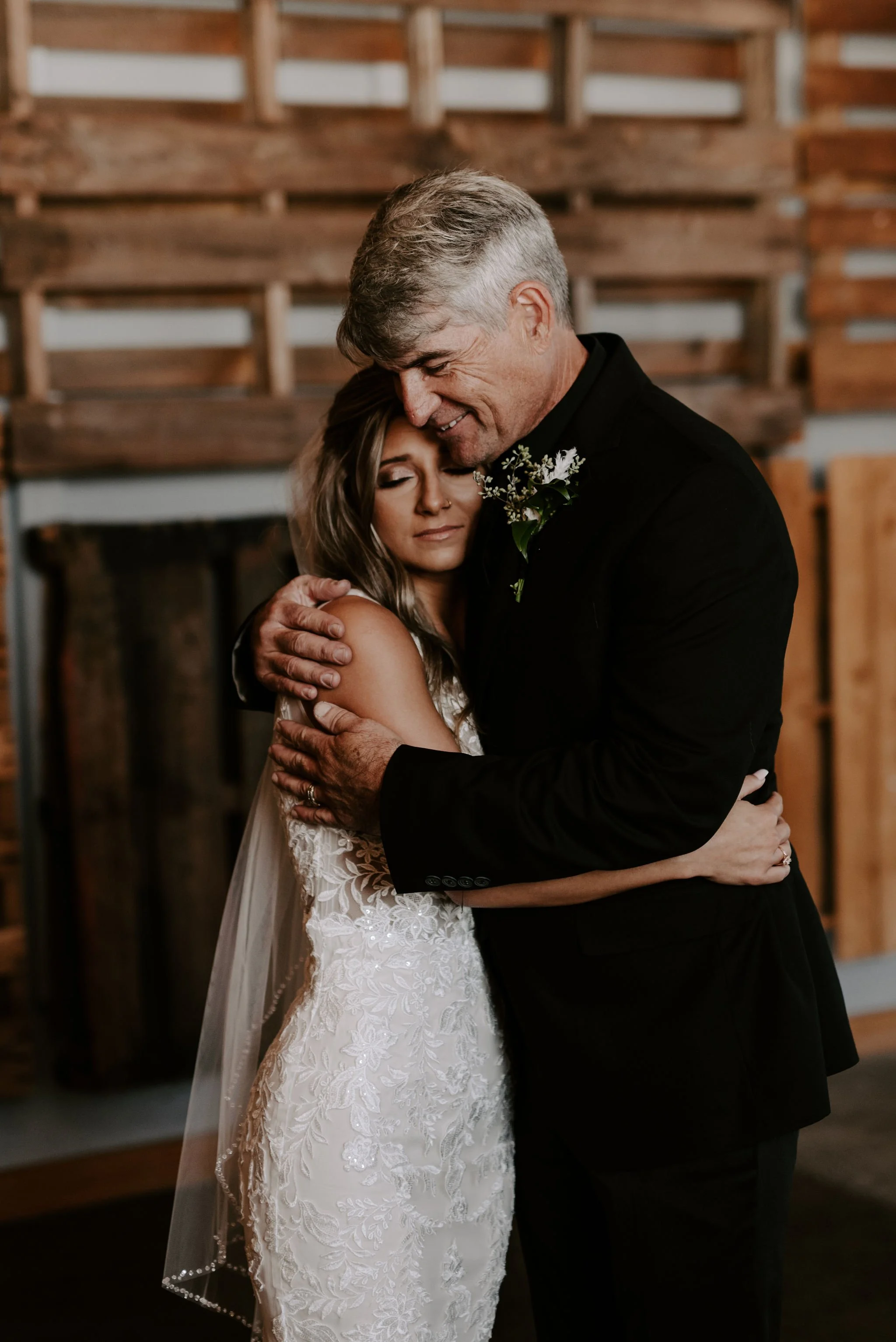 A bride embracing an older man, possibly her father, at a wedding in a cozy, rustic setting with wooden walls.