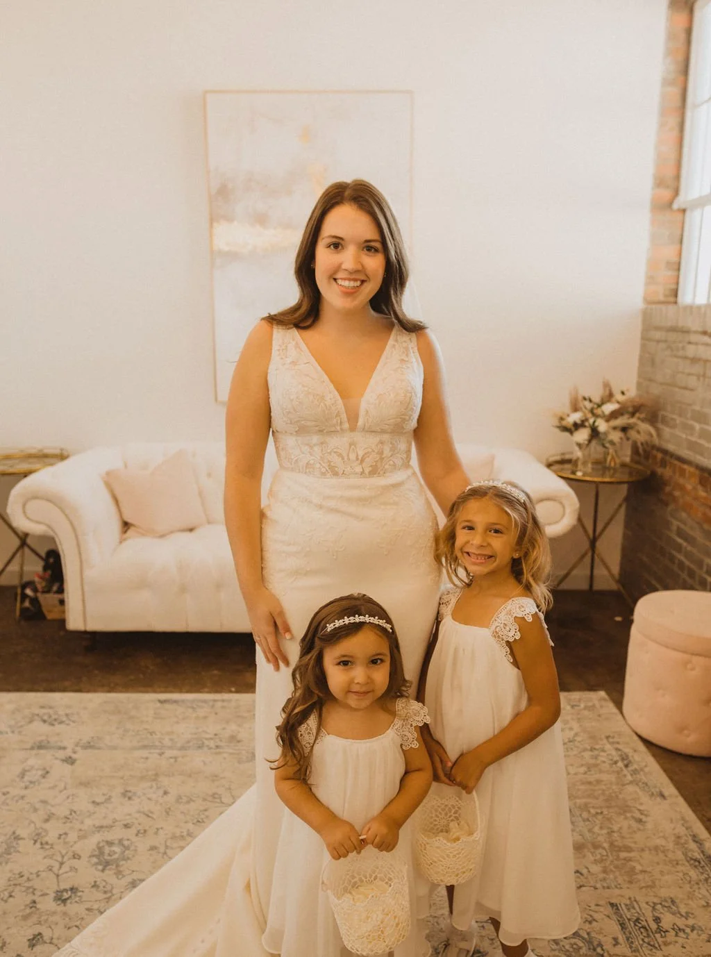 A woman in a wedding dress standing with two young girls also in white dresses, inside a cozy room with a white couch, abstract art on the wall, and a brick wall near a window.