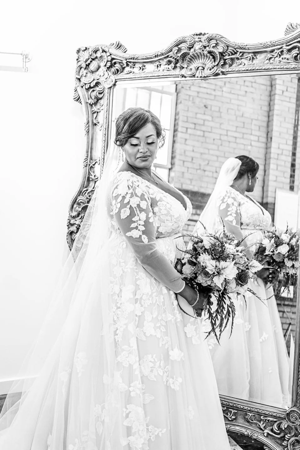A bride in a wedding gown with floral lace details, holding a bouquet of flowers, standing in front of an ornate mirror, with her reflection visible.