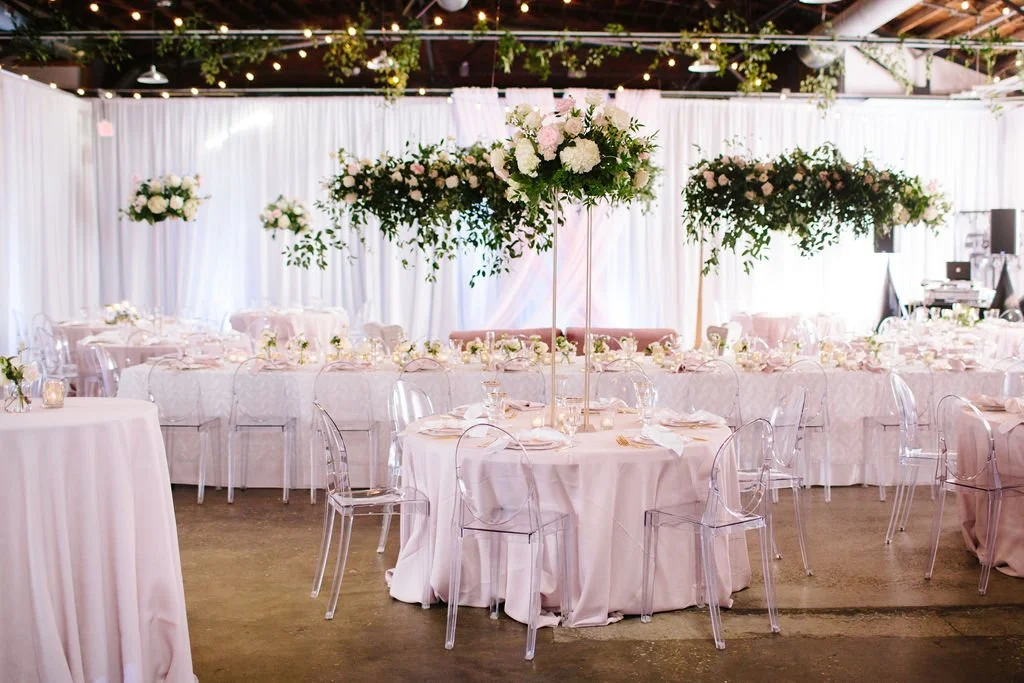 Elegant event space decorated with floral centerpieces, white drapery, and transparent chairs, set up for a formal occasion or wedding reception.