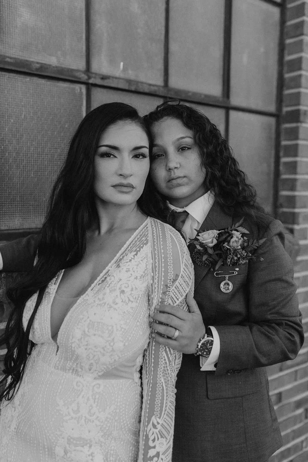Black and white photo of two women, one in a wedding dress and the other in a suit with a flower pin, standing close together against a brick and panel wall. The woman in the wedding dress has long dark hair, and the woman in the suit has curly hair 