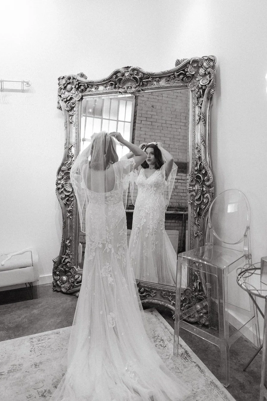 A woman in a wedding dress with a veil looks at herself in an ornate, large mirror in a bright room with a brick wall and window, preparing for her wedding.