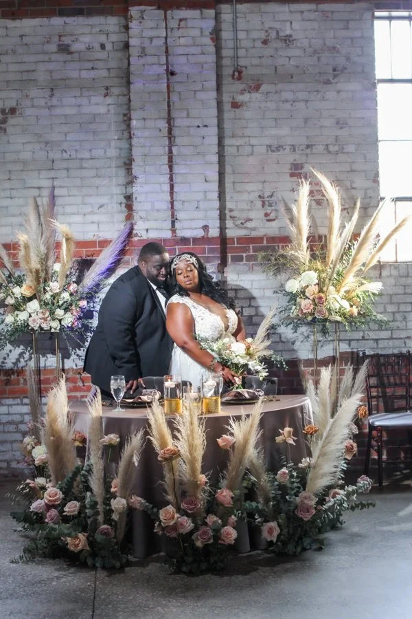 A couple is at a wedding reception, standing behind a decorated table with floral arrangements, glasses, and drinks, inside a rustic industrial venue with brick walls and large windows.